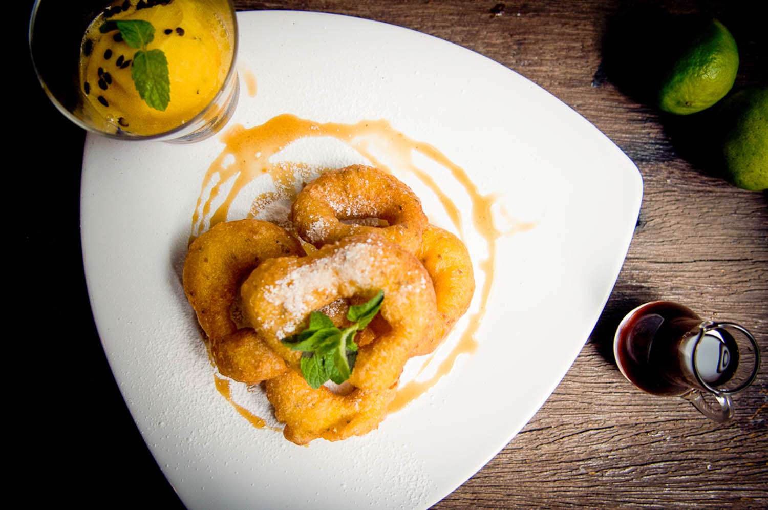An overhead view of Peruvian picarones on a platter drizzled with sauce and garnished with fresh herbs. Next to a glass, a small pitcher of syrup, and two limes.