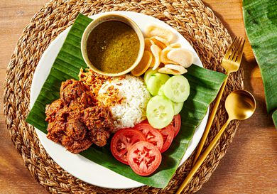 Plate of beef Rendang with rice, cucumbers, tomatoes and crisps on a banana leaf, with a woven placemat and gold cutlery