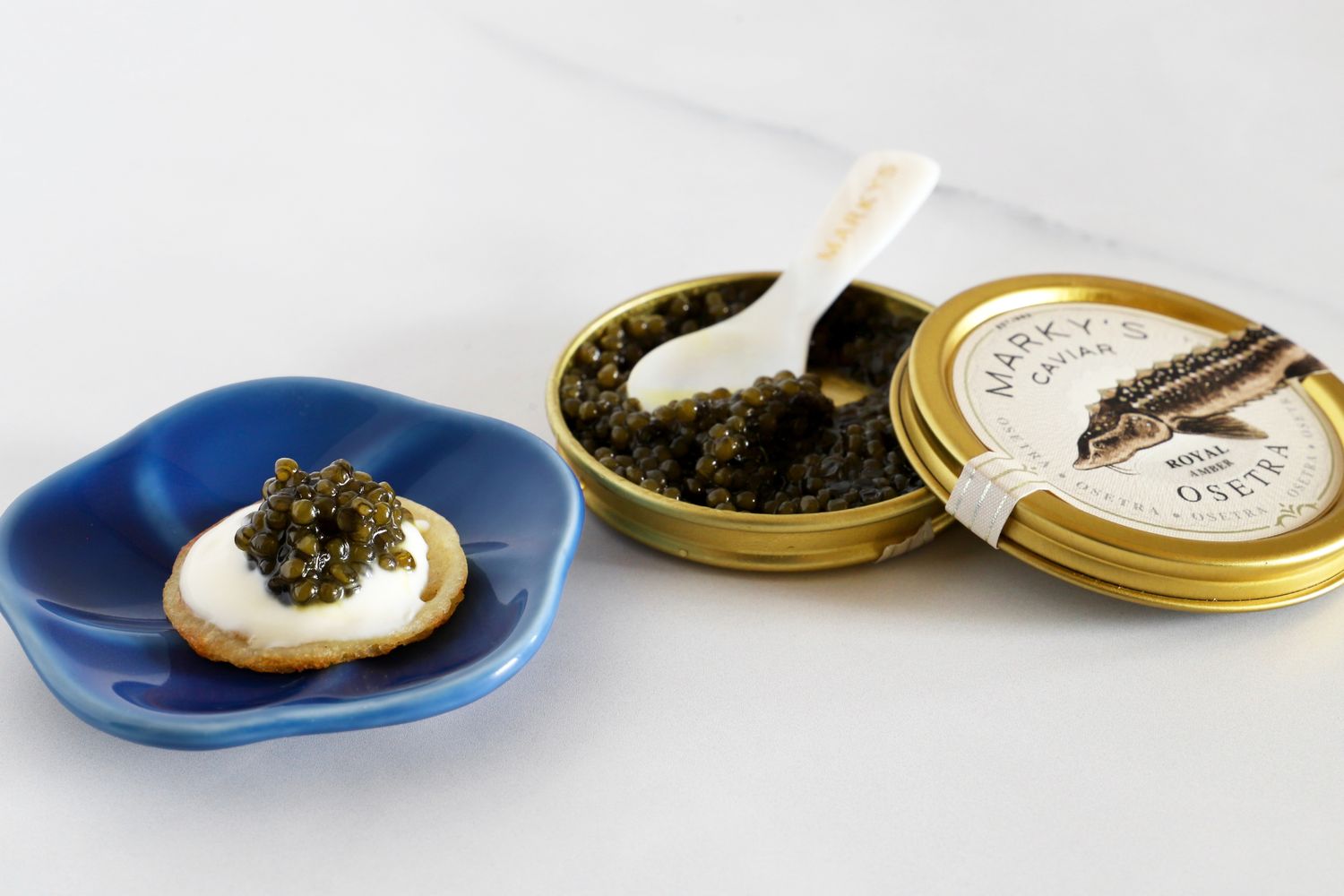 a tin of Marky's osetra caviar next to a blini in a blue dish topped with crème fraîche and a scoop of caviar