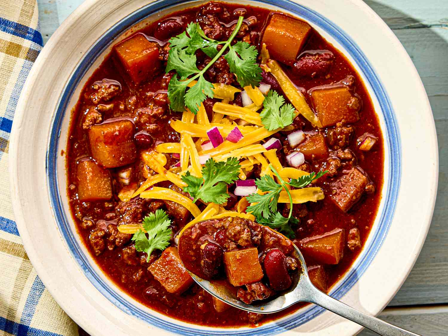 A bowl of turkey chili with squash beans cheese onions and cilantro garnish spoon served alongside