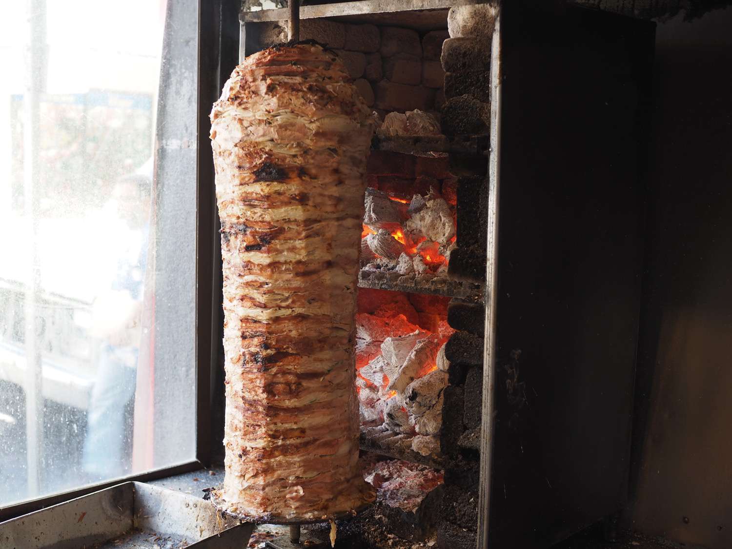 A trompo (vertical spit) loaded with the pork for tacos árabes rotates next to a shelf stacked with hot coals.