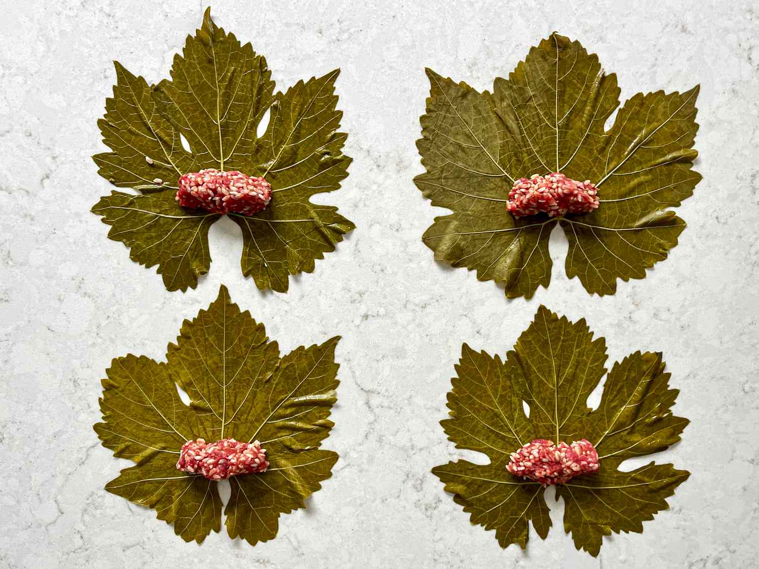 Four grape leaves arranged on a grey counter with small logs of meat mixture placed at the base of each left