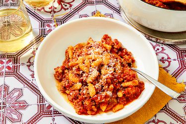 Overhead view of Malloreddus alla Campidanese in a white bowl on a titled background with pink floruishes surrounded by the pot and two wine glasses