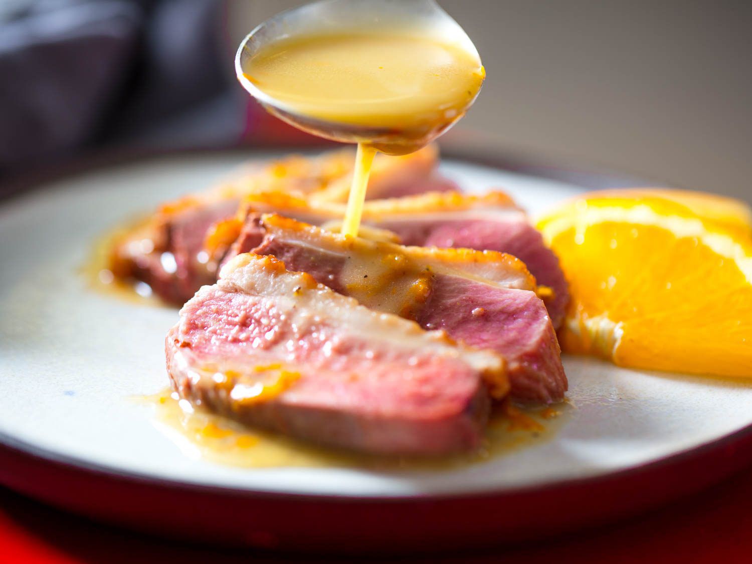 Ladle pouring pan sauce over medium-rare seared duck breast plated with orange slices