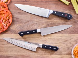 Three chef's knives on a wooden countertop with sliced tomatoes nearby.