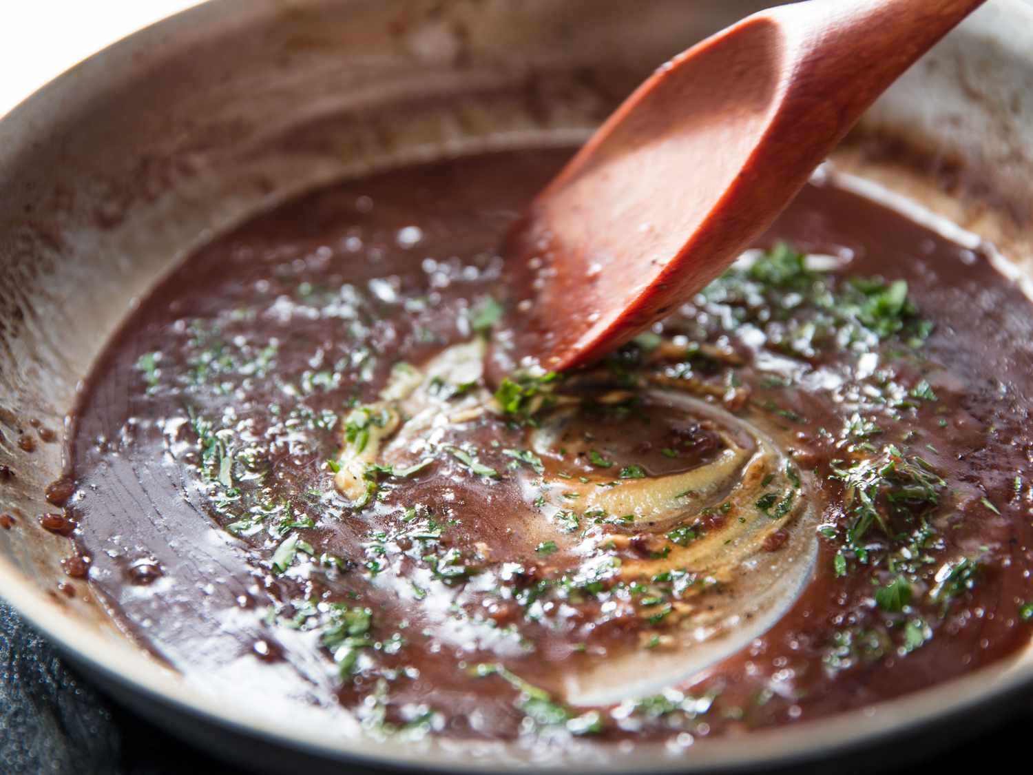 A pan sauce made with red wine and fresh herbs is brought together in a skillet with a flat-edged wooden spoon.