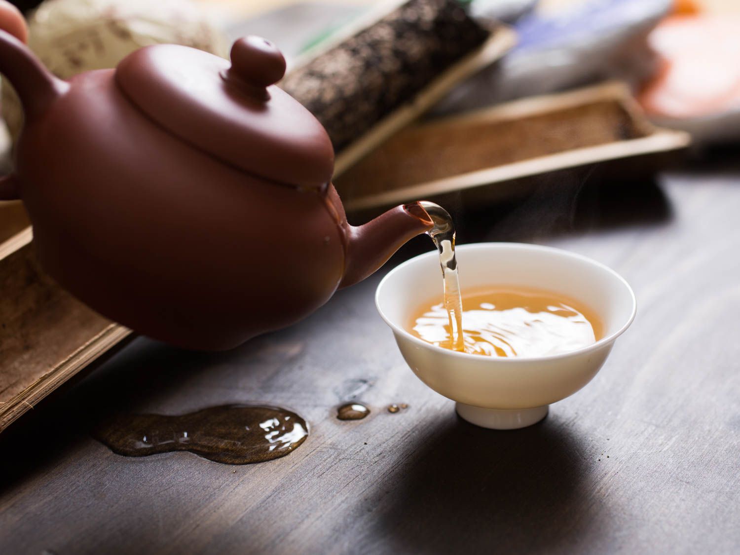 Tea being poured into a small white cup from a teapot with spills and tea ware visible