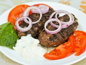 Greek bifteki on a plate with tomato slices, red onion slices, mint leaves, and tzatziki.