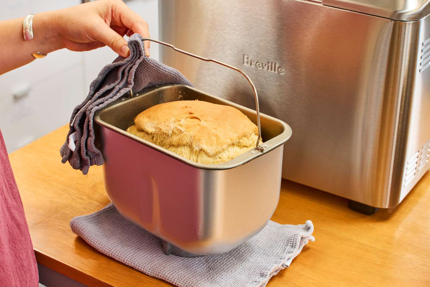 A close-up of a hand using a towel to hold a metal basket in front of the Breville Custom Loaf Bread Maker containing baked bread