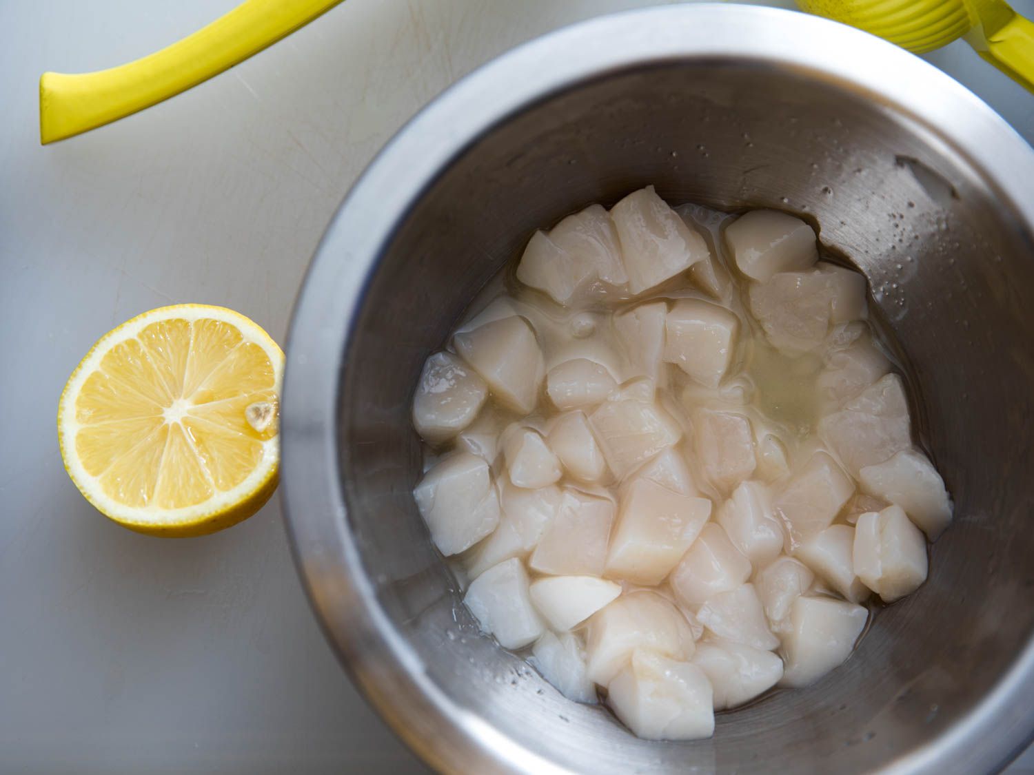 Uncooked scallops marinating in a stainless bowl filled with lemon juice, a half lemon and juicer on the side.