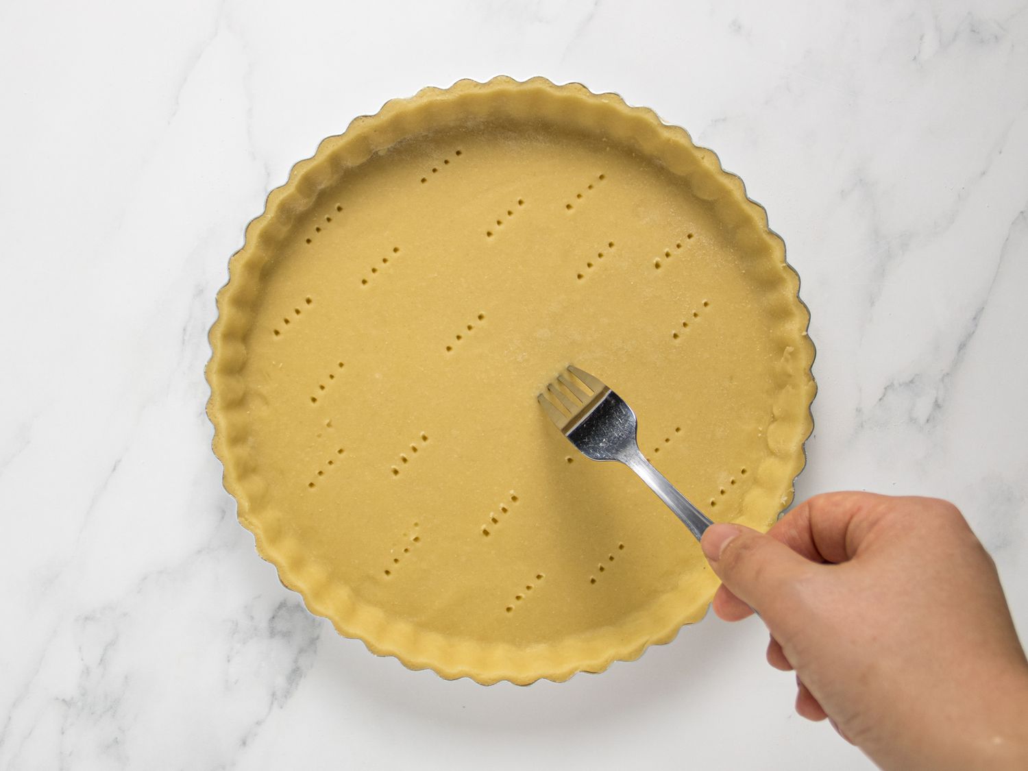 Hand with fork poking holes into an uncooked pate brisee on a marble surface 