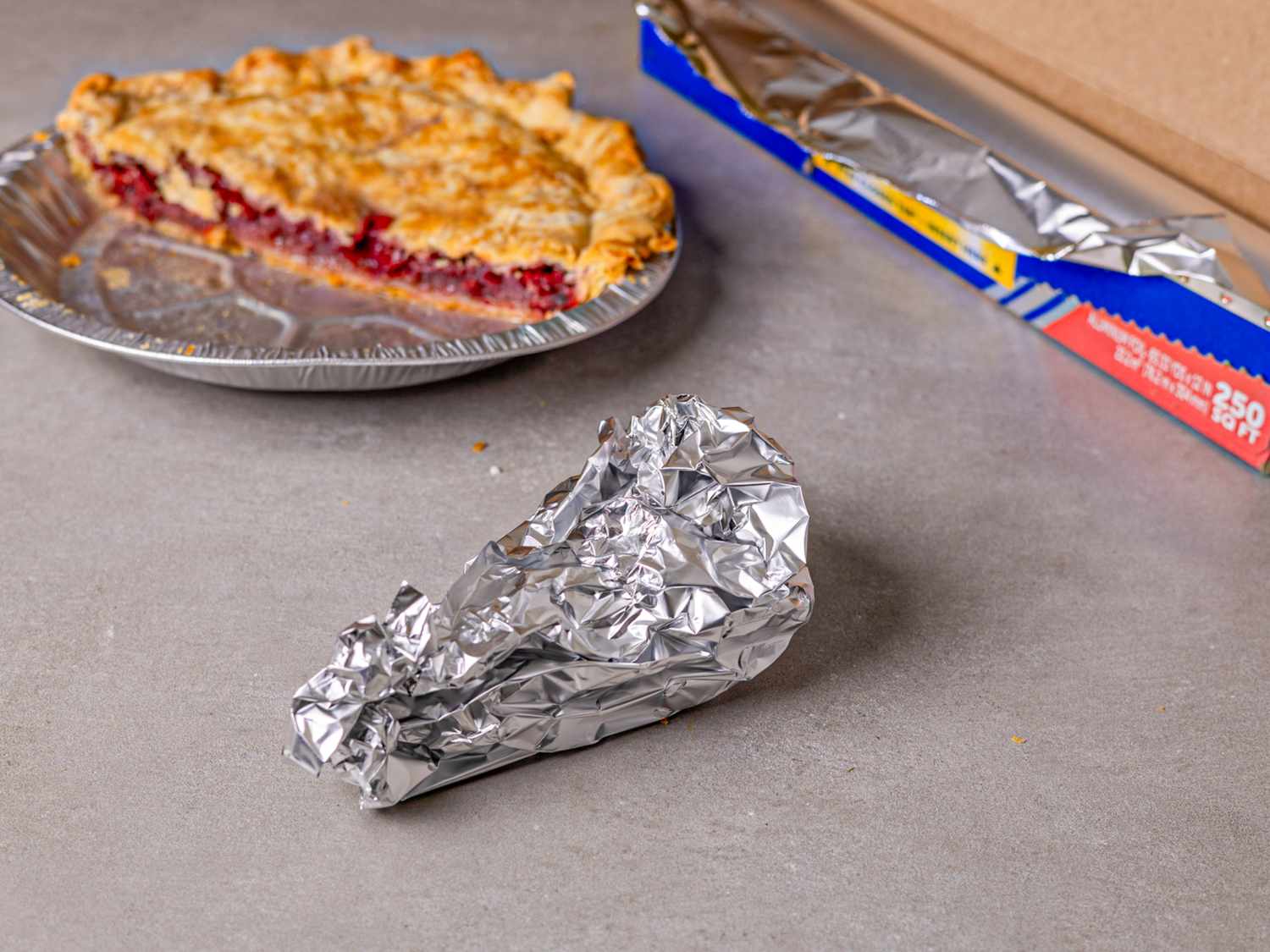A partially eaten pie and an aluminum foil wrap on a gray surface with a foil box in the background