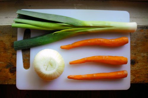 an onion, a leek, and three peeled carrots on a white plastic cutting board