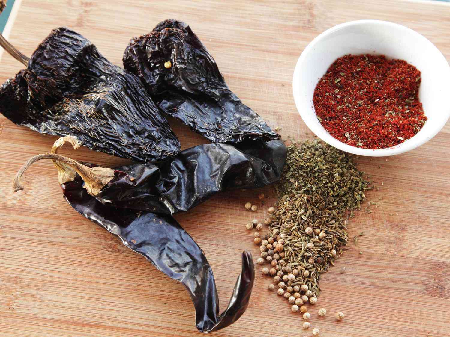 Large dried chilies, spices, and a bowl of chili powder on wooden cutting board