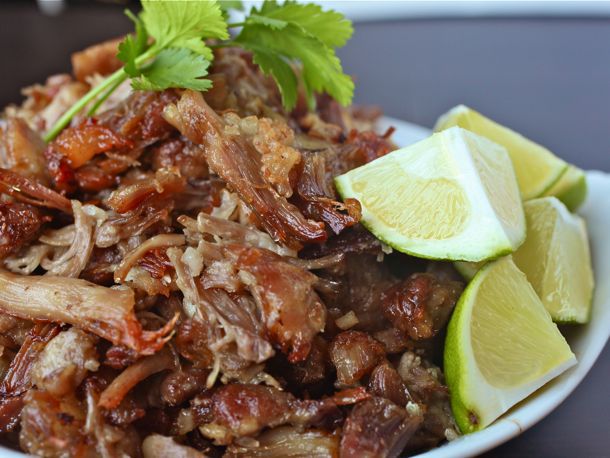 Bowl of braised and crisped carnitas topped with lime wedges and cilantro.