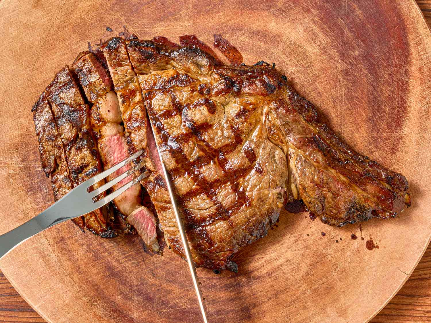 A cooked steak on a wooden board being sliced and held with a fork