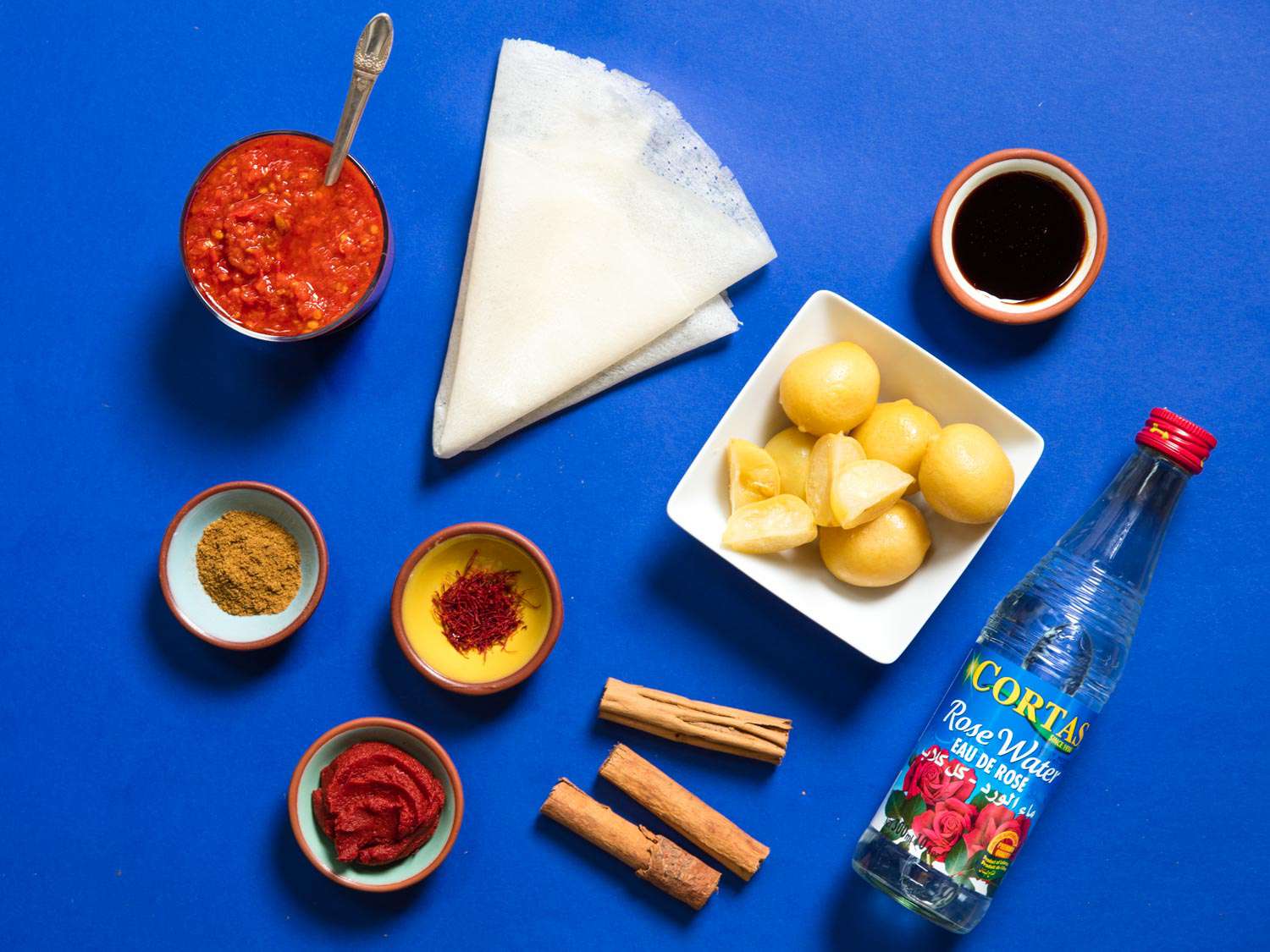 A collection of ingredients used in Moroccan cooking, including preserved lemon, rose water, cinnamon, saffron, Ras el Hanout, harissa, and a sheet of warqa pastry 