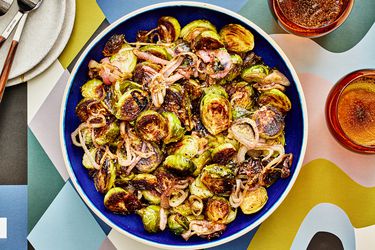 Roasted Brussels sprouts and shallots with a balsamic glaze in a bowl, with glasses of sparkling water, some smaller plates and a colorfully printed table top