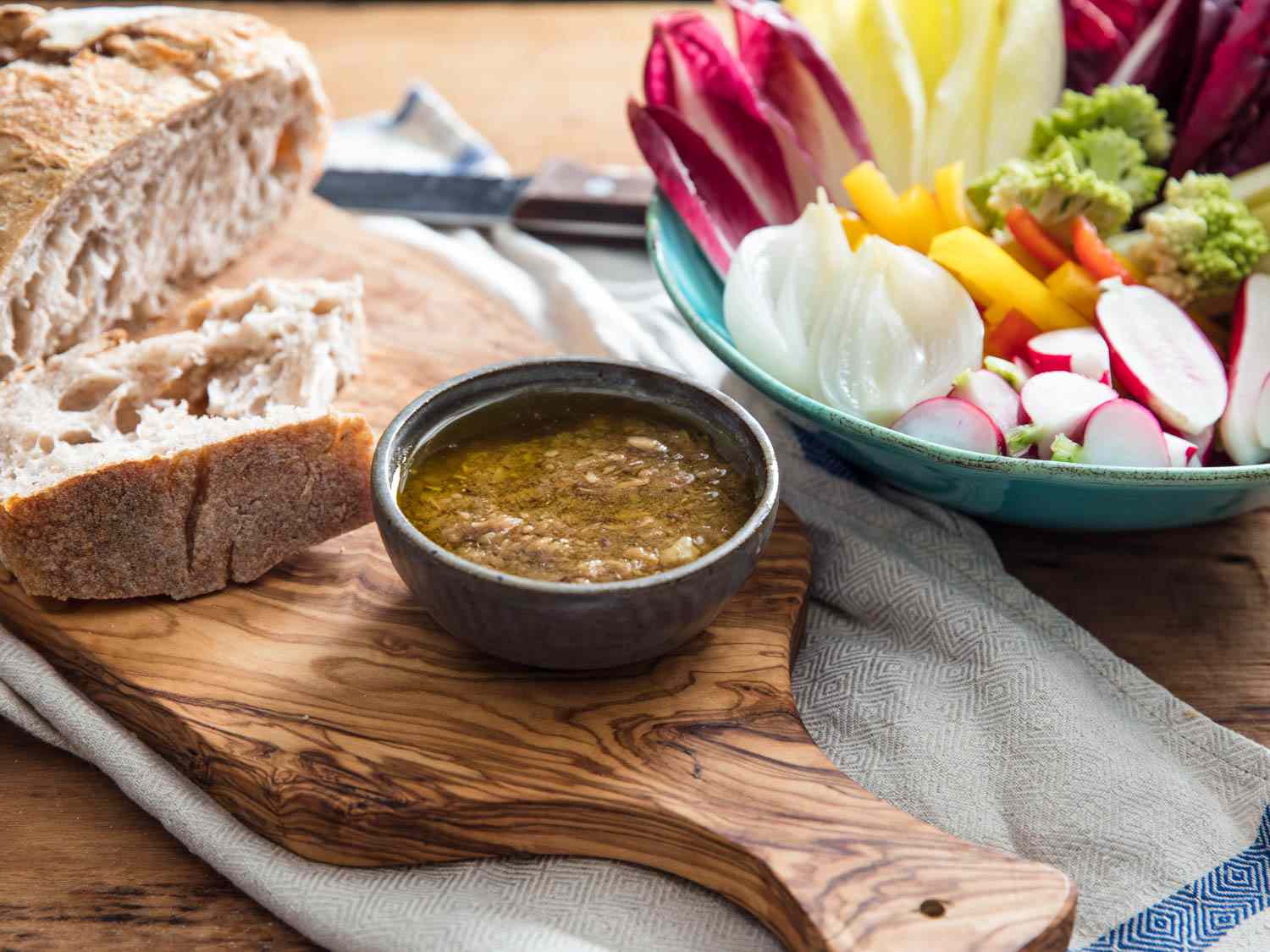 A wooden platter with a small bowl of bagna cauda anchovy dipping sauce, fresh bread, and crudite.