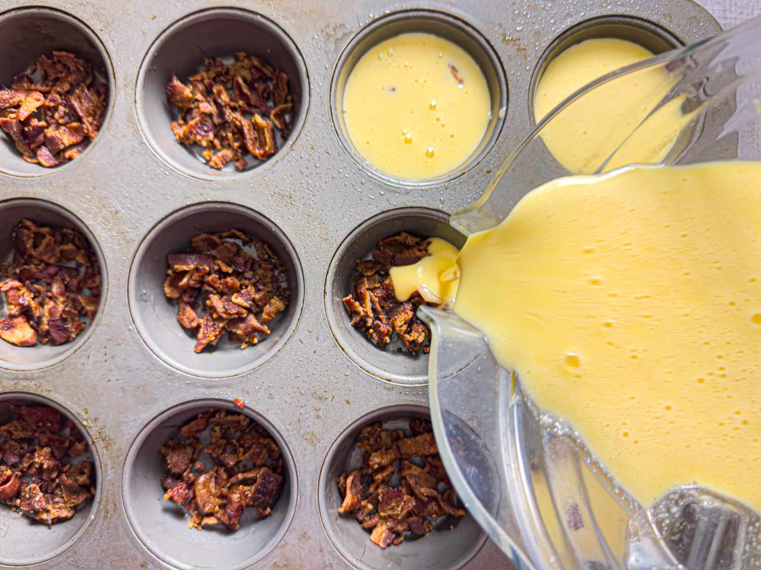 Egg mixture being poured into a muffin tin containing bacon bits