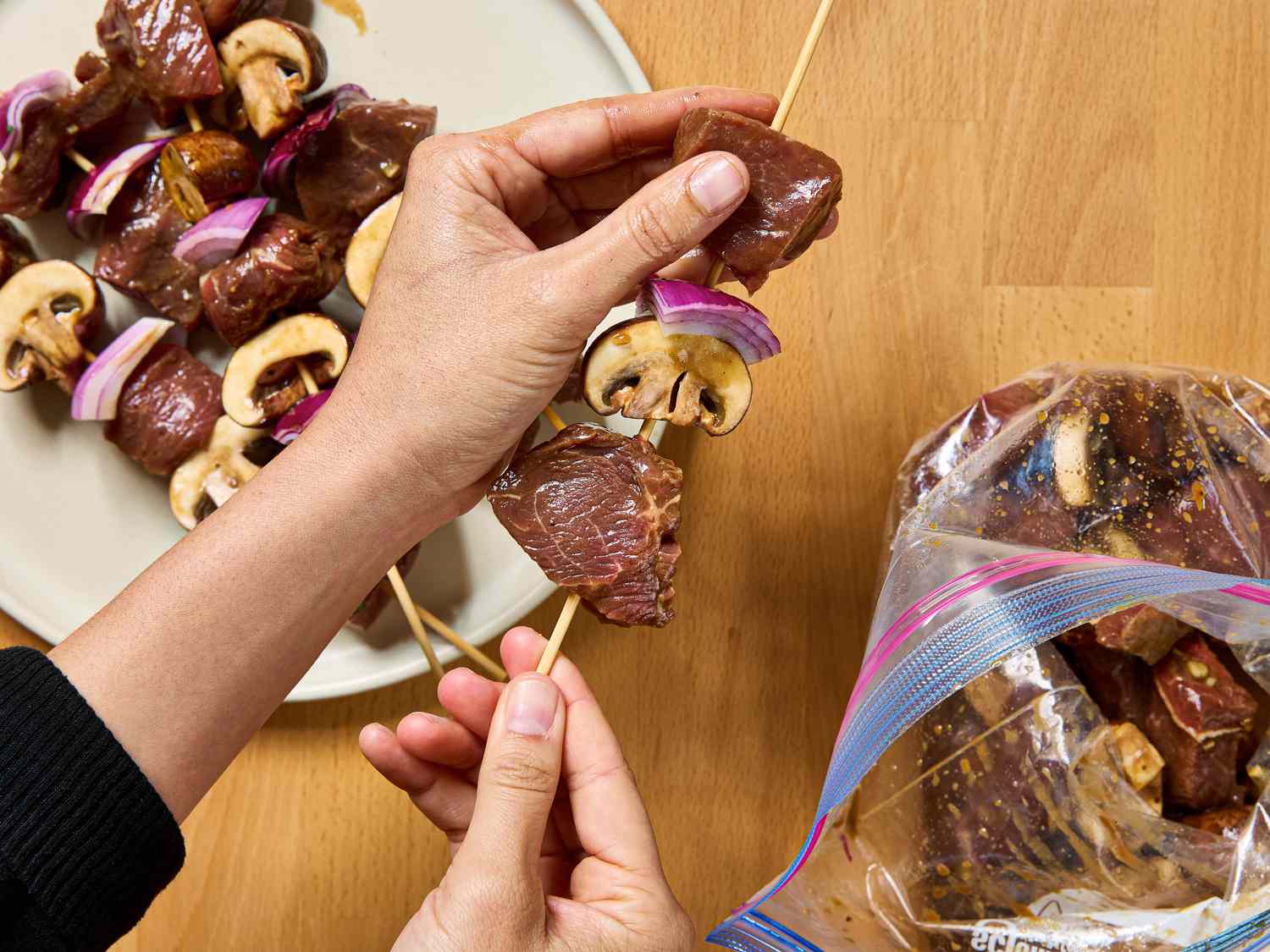 Person assembling kebabs with steak, mushrooms, and onions on wooden skewers, ingredients on a plate and marinade in a bag nearby