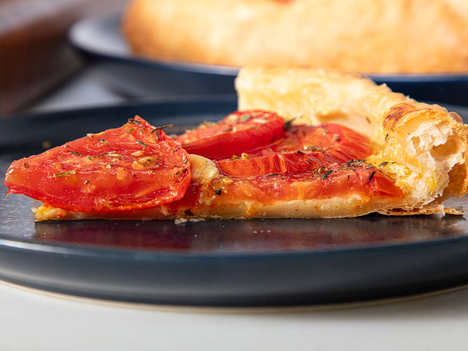 A slice of tomato tart served on a plate featuring puff pastry and seasoned tomato slices on top