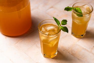 Two tall glasses of Arnold Palmer, each with a sprig of mint. In the top left corner of the image is a pitcher holding more Arnold Palmer.