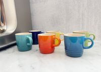Six Le Creuset espresso cups in different colors on a marble countertop.