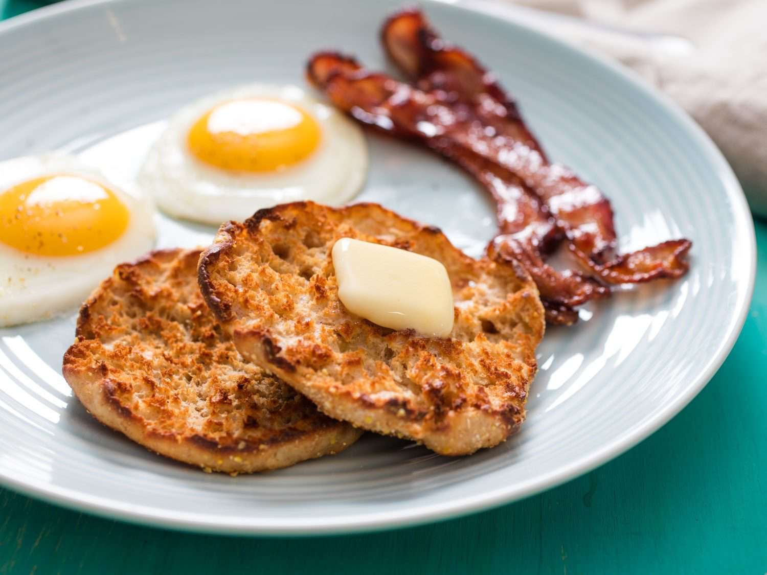 A toasted English muffin with butter, on a plate with eggs and bacon.