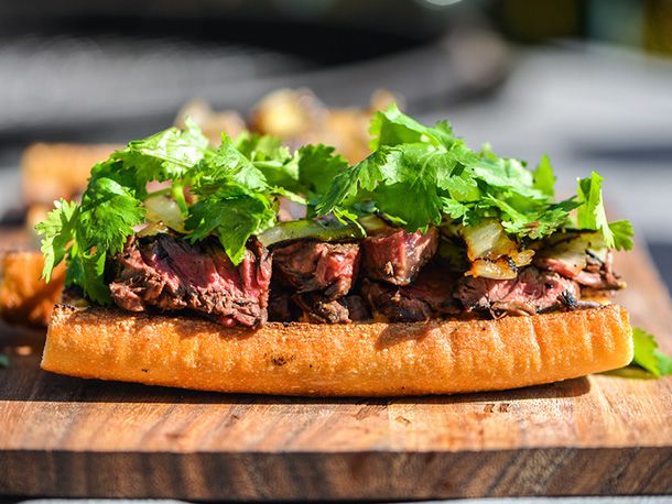 Sliced grilled steak piled onto the bottom half of a baguette, topped with grilled onion and fresh cilantro on wooden board 
