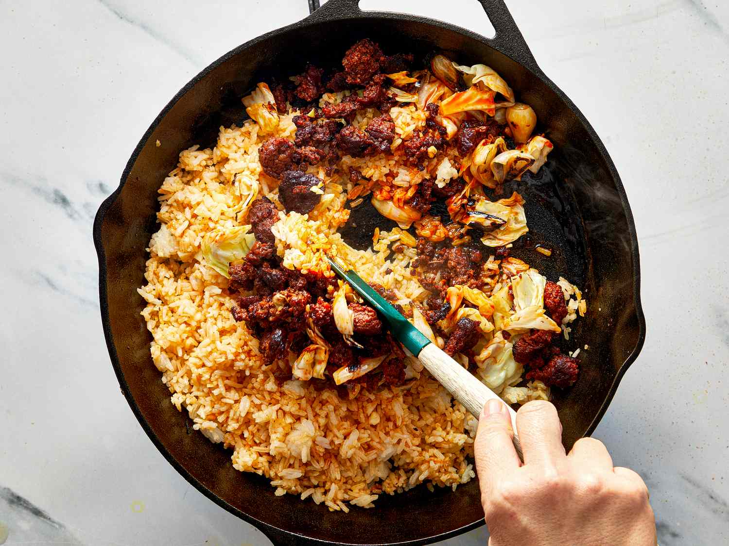 A skillet with Mexicaninspired fried rice being stirred by a hand using a utensil featuring rice vegetables and chorizo