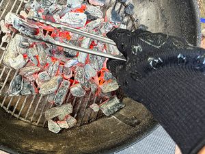 A hand in a heatresistant glove holding metal cooking tongs over smoldering charcoal in a grill