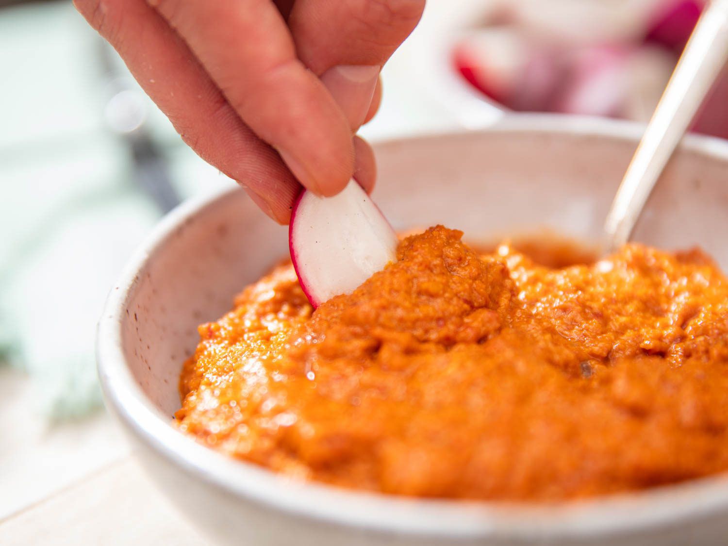 Dipping a raw radish in vibrant red-orange romesco sauce.