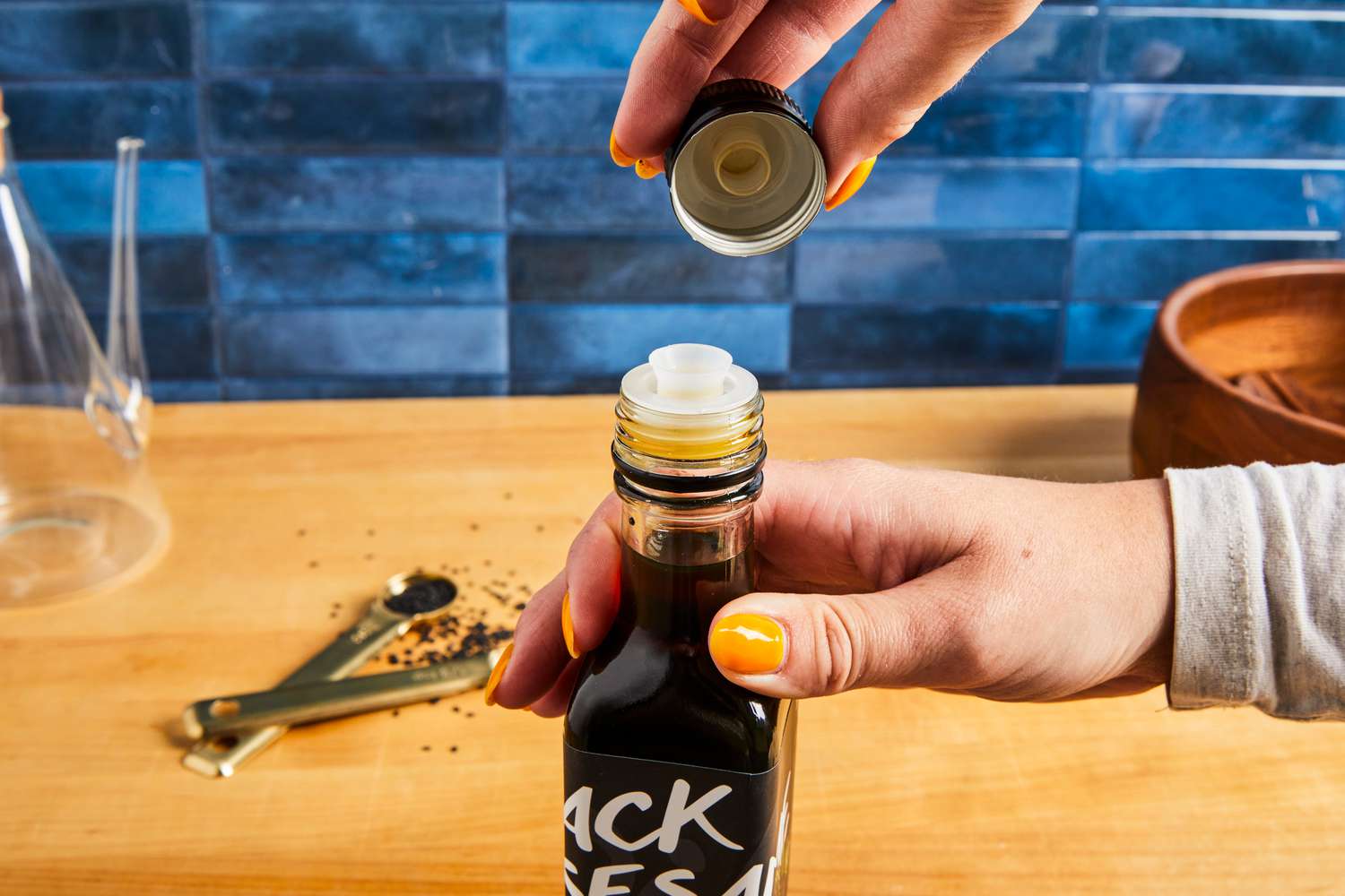 Hand holding a bottle of black sesame oil with the lid being removed, kitchen surface with spilled seeds in the background