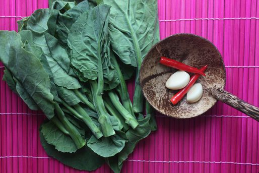 Overhead view of a bunch of Chinese broccoli set on a fuchsia bamboo mat. A coconut-wood ladle is set nearby, cradling red chiles and garlic cloves.