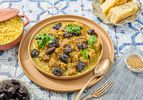 Lamb and prune Tagine with side of couscous, prunes, bread and cutlery on a blue tile surface 