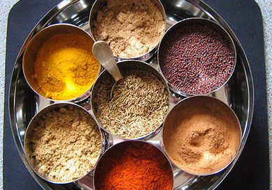 A collection of seven dry spices in a masala dabba, a large tin with smaller tins inside.