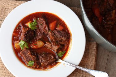 A white serving bowl of goulash sits next to a Dutch oven.