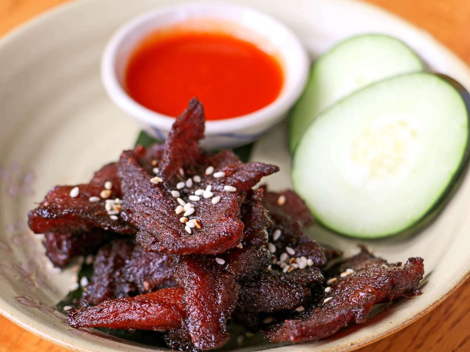 A stoneware plate of dark-red sun-dried pork (muu haeng) next to sliced cucumber and a small bowl of dipping sauce.