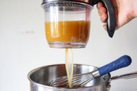 A handheld kitchen tool dispensing liquid from a container into a saucepan containing a whisk