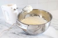 An electric handheld mixer beside an All-Clad mixing bowl containing partially whipped cream