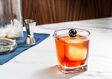 A Boulevardier cocktail in a cocktail glass with one large cube of ice, garnished with a cherry on a cocktail pick.
