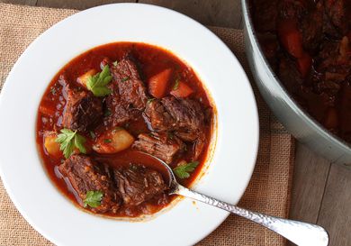 A white serving bowl of goulash sits next to a Dutch oven.