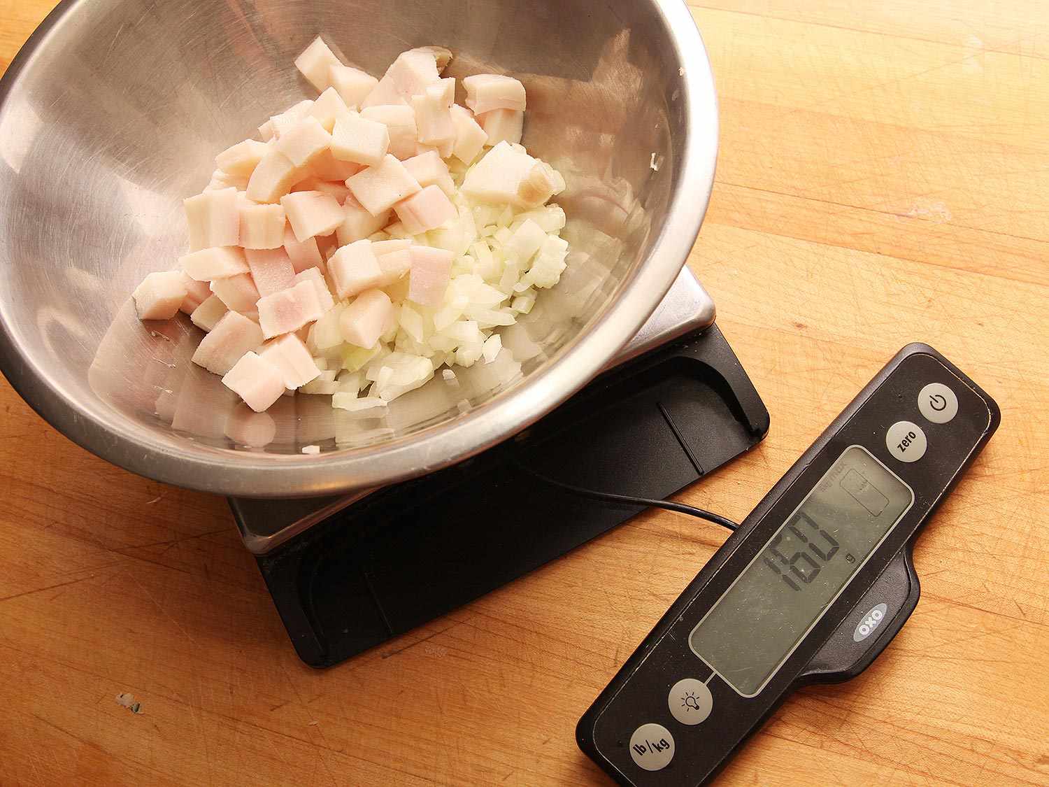 A mixing bowl of cubed pork fat and chopped onion is weighed on a digital scale.