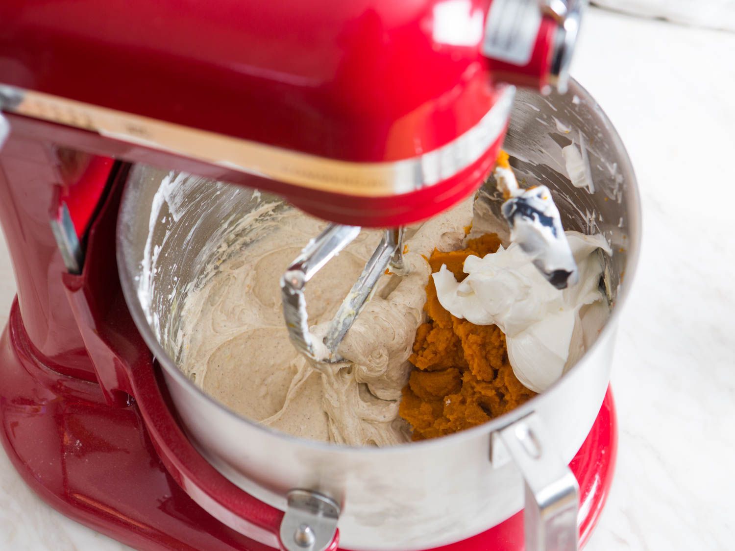 Adding pumpkin puree and sour cream to stand mixer bowl of cheesecake mixture to make pumpkin cheesecake.