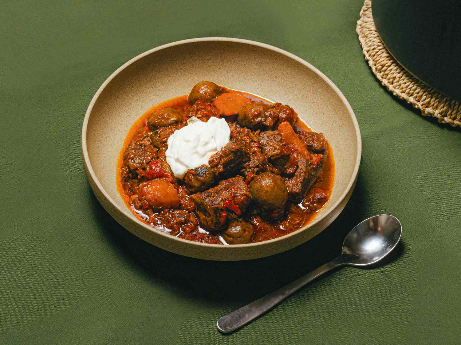 A bowl of Spanish beef stew topped with sour cream served on a green tablecloth with a spoon nearby