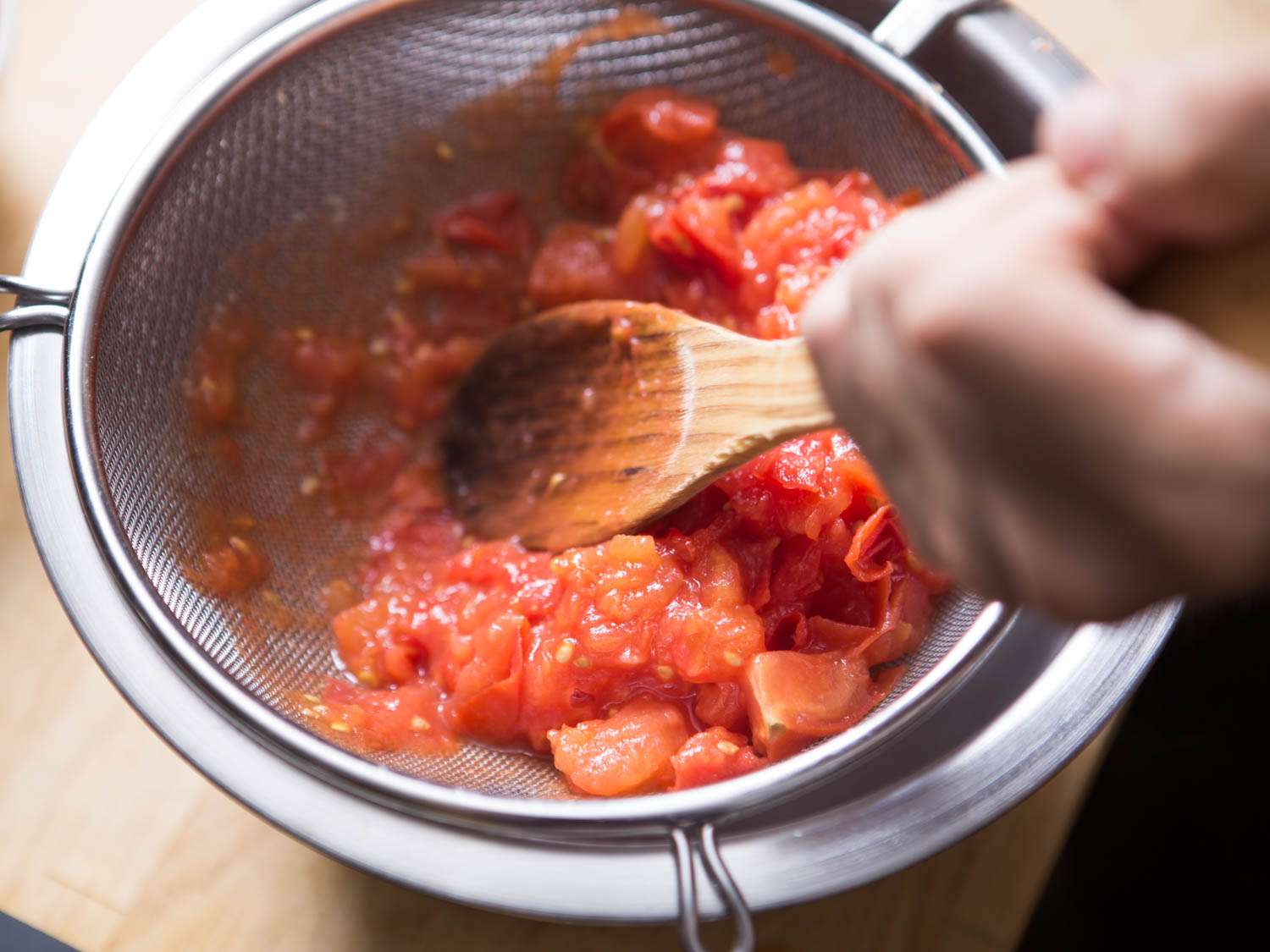 Simmered tomatoes being pressed and scraped through a fine mesh strainer.