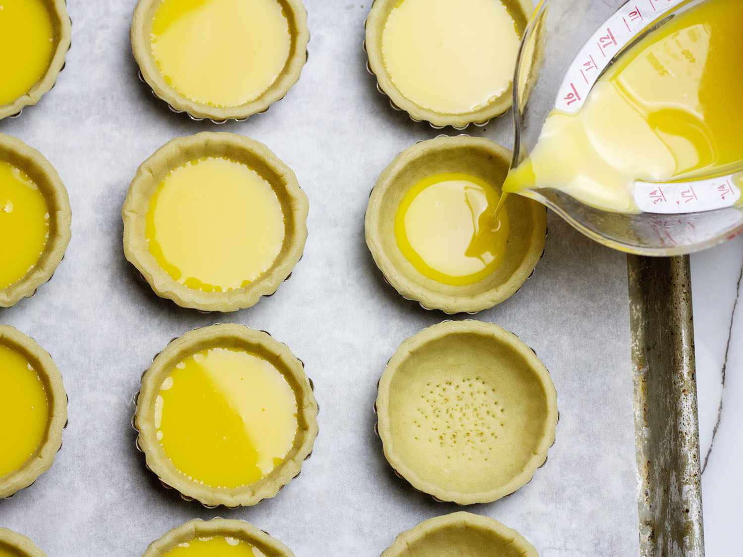 Custard being poured into Hong Kong egg custard tart shells on a parchment lined baking sheet