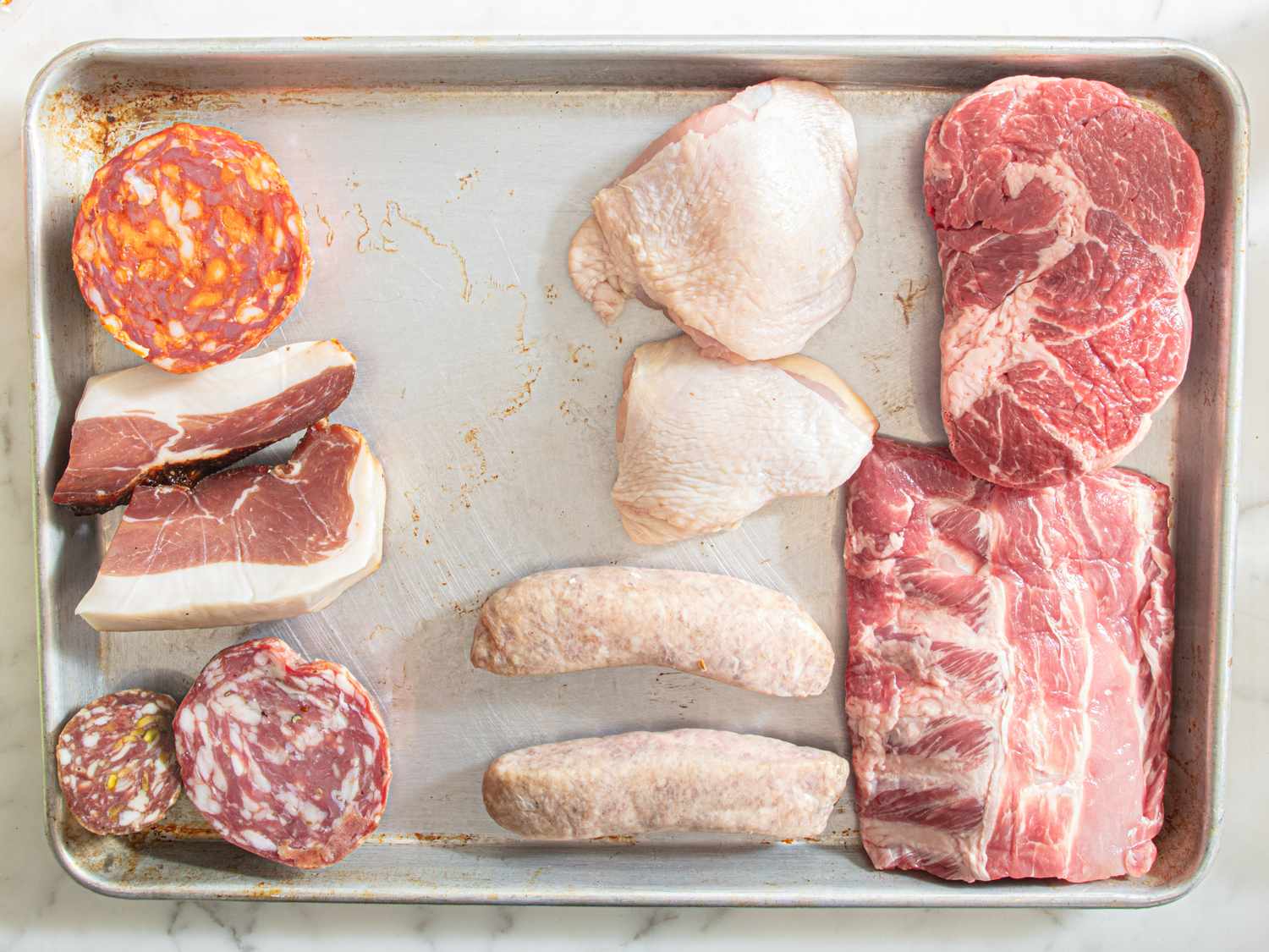 A metal tray displaying an assortment of raw meats including sausages sliced cured meats chicken pieces and beef cuts