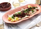 A dish of grilled fish topped with olive and tomato compote served on a table with lemon wedges and parsley garnish